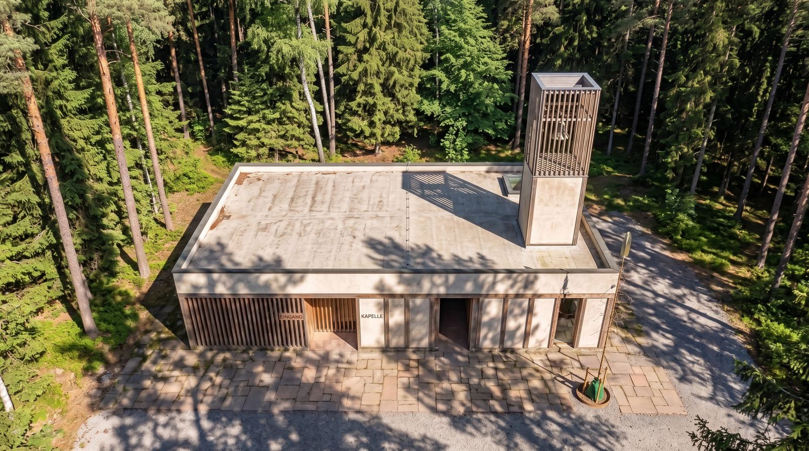 Aerial view of the flat-roofed concrete chapel with its vertical timber bell tower casting a shadow across the roof, surrounded by dense green forest.