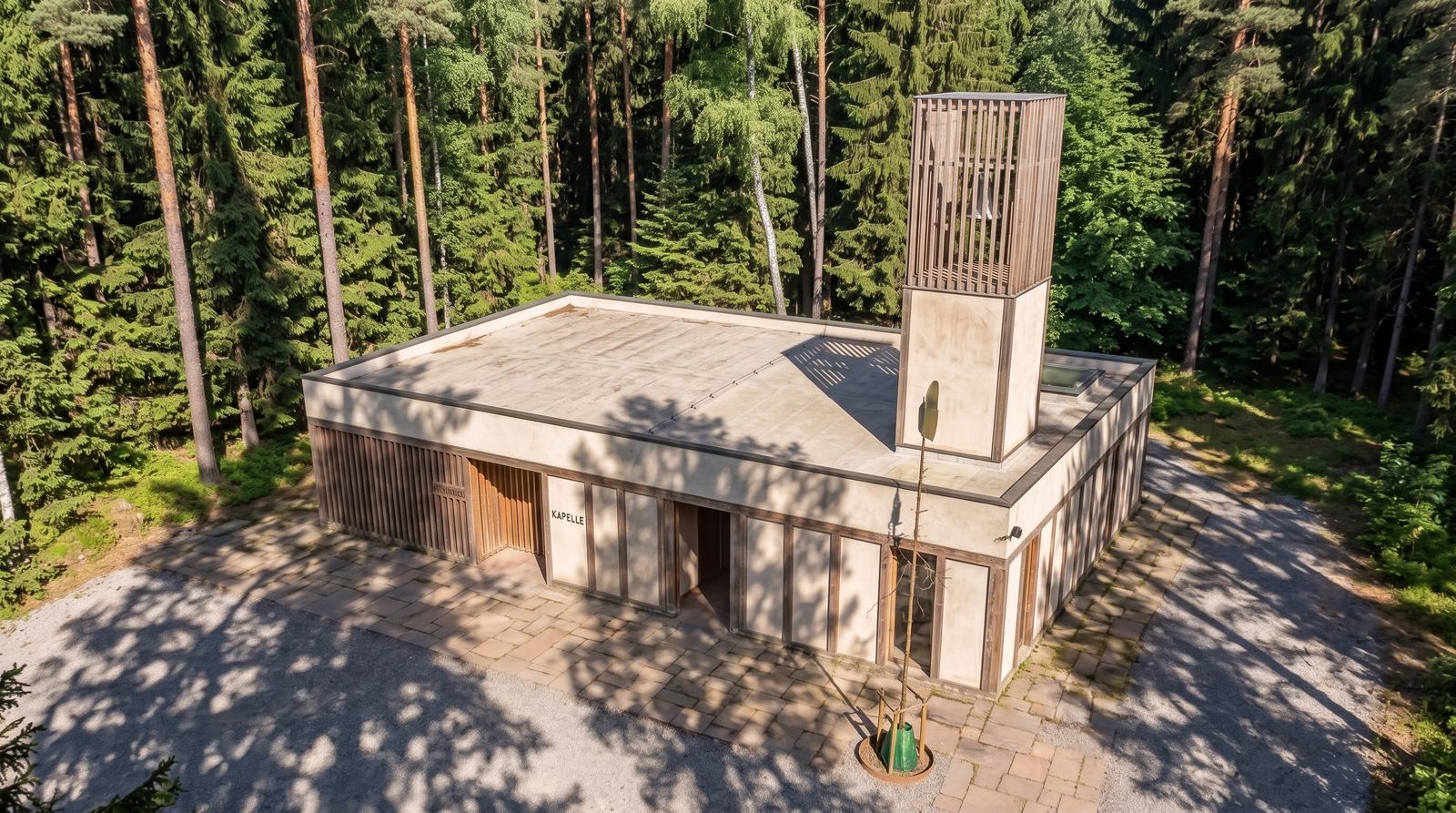 An elevated drone perspective of the rectangular concrete chapel showing its flat roof, vertical timber bell tower, stone-paved courtyard, and surrounding dense forest canopy.