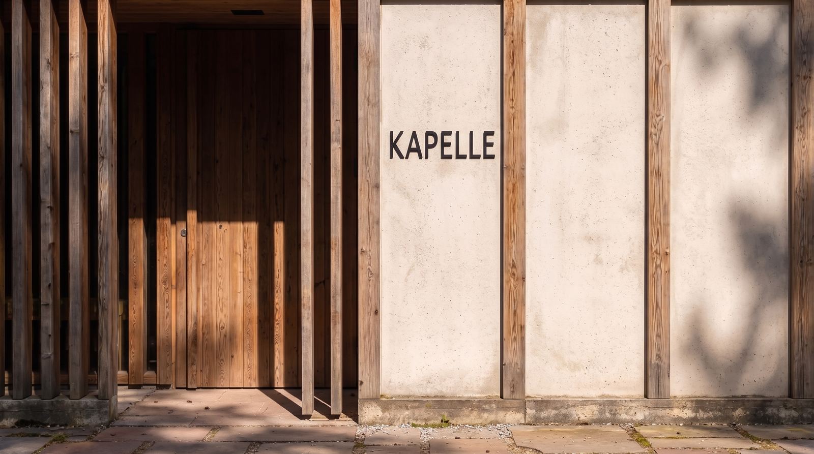 Close-up of the textured concrete wall featuring the inscribed text "KAPELLE", framed by vertical timber columns and a recessed wooden entrance with casting shadows.
